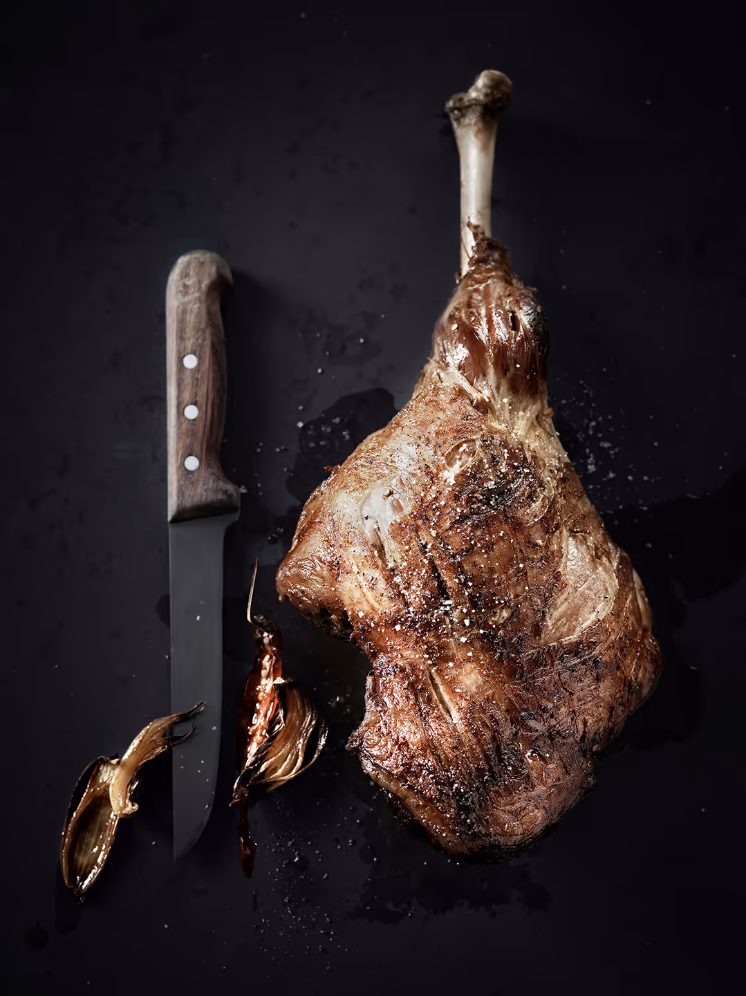 Gegrilltes Lammkeulenstück mit Knochen, daneben ein Messer mit Holzgriff und gebratene Knoblauchzehen auf dunklem Untergrund.
