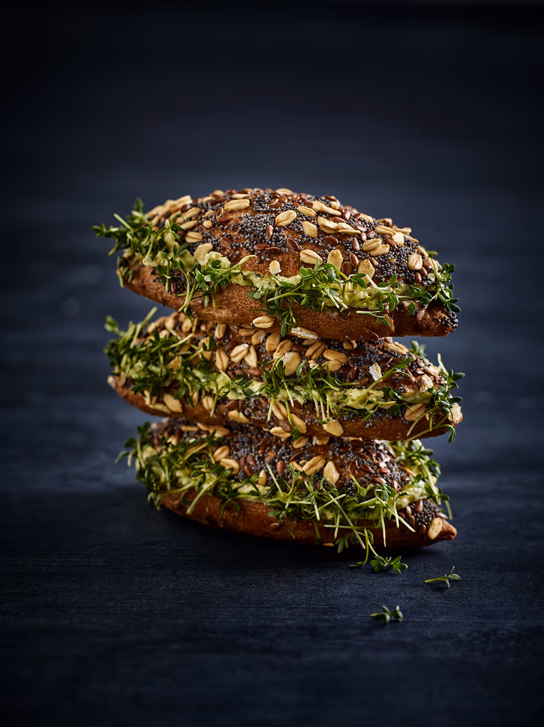 Drei mit Körnern bestreute Brötchen, gefüllt mit grünem Aufstrich und Kresse, gestapelt auf dunklem Untergrund.