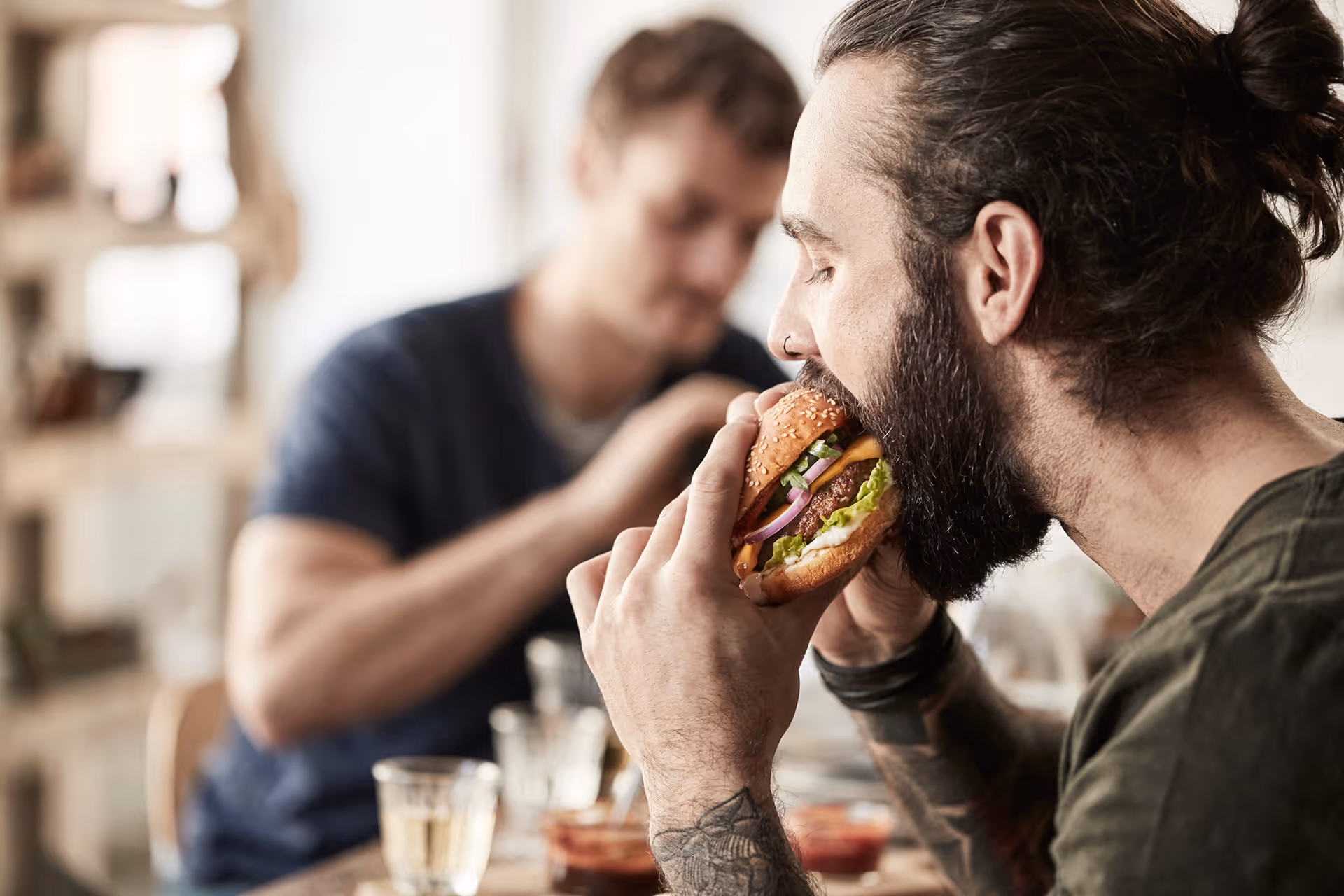Ein tätowierter Mann mit Pferdeschwanz beißt in einen Burger mit Salat, Käse und Zwiebeln, während ein weiterer Mann im Hintergrund isst.