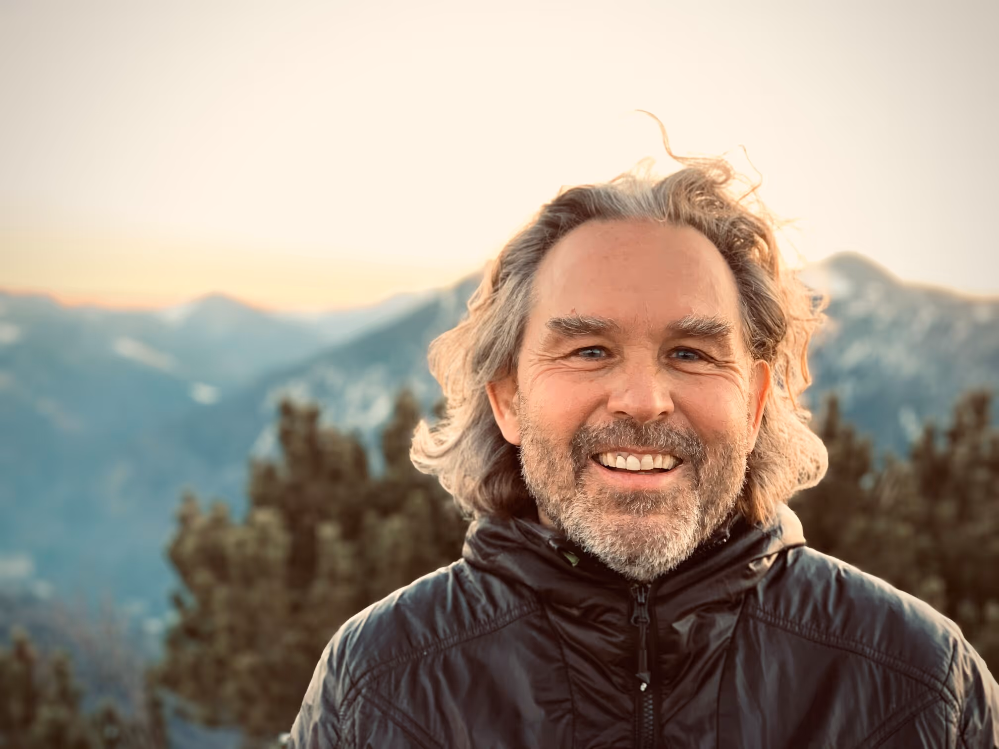 Ein lächelnder Mann mit grauem Haar und Bart, der eine schwarze Jacke trägt, vor einer Berglandschaft bei Sonnenuntergang.