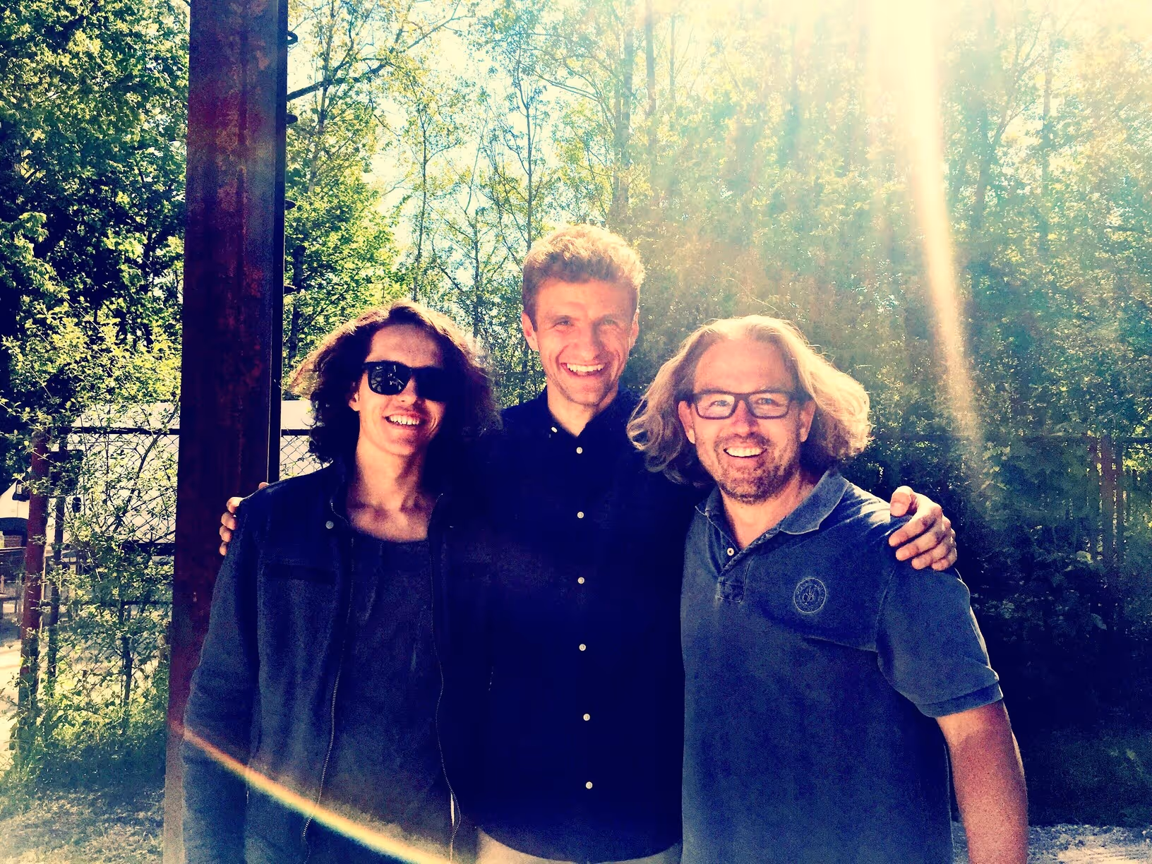 Drei lächelnde Männer stehen im Freien in der Sonne vor Bäumen, einer trägt eine Sonnenbrille.