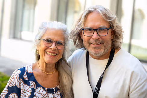 Mann mit längeren blonden Haaren und Outdoor-Bekleidung sitzt auf einem Holzbrett vor felsigem und grasbewachsenem Gelände, neben ihm Essen und zwei Wasserflaschen.