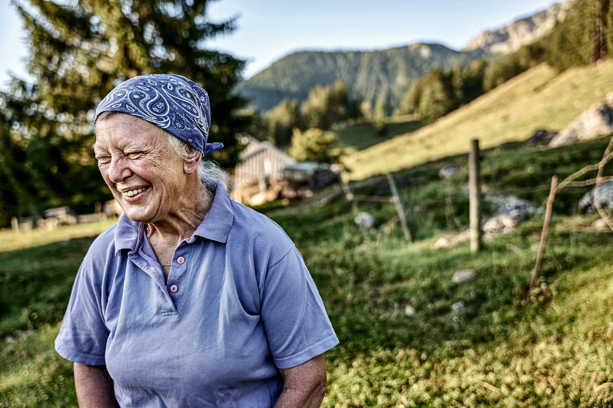 Lächelnde ältere Frau mit Kopftuch auf Almwiese vor bewaldeten Bergen.