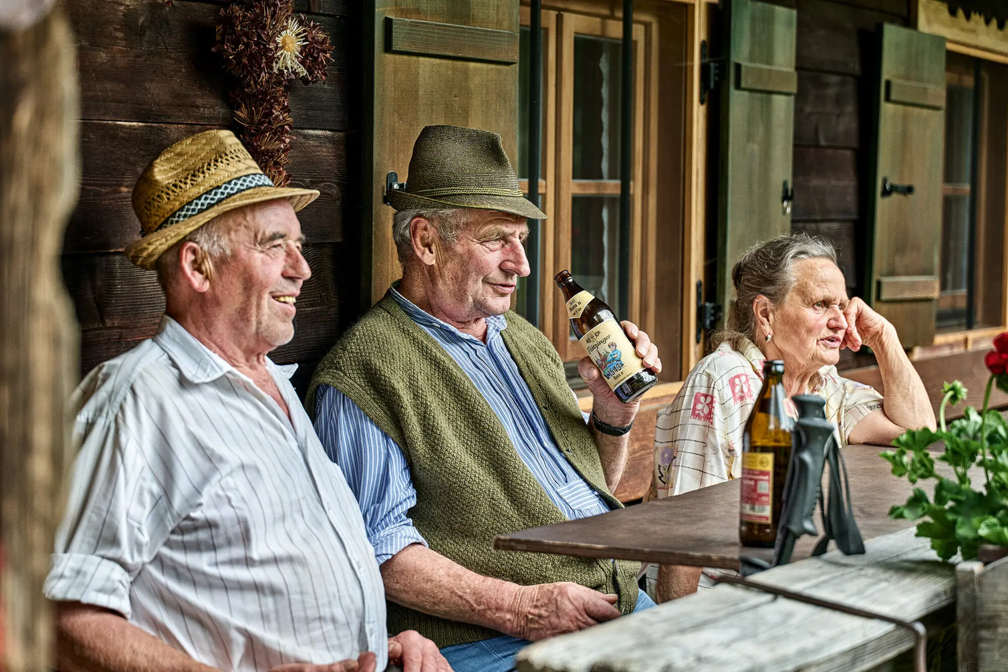 Drei ältere Personen mit Bier auf rustikaler Almhütten-Terrasse.