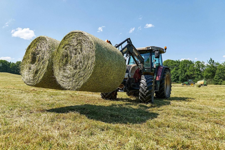 Traktor mit Frontlader hebt zwei große Heuballen auf Feld.
