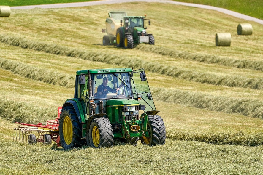 Zwei Traktoren arbeiten in hügeligem Feld auf Heuschwaden.