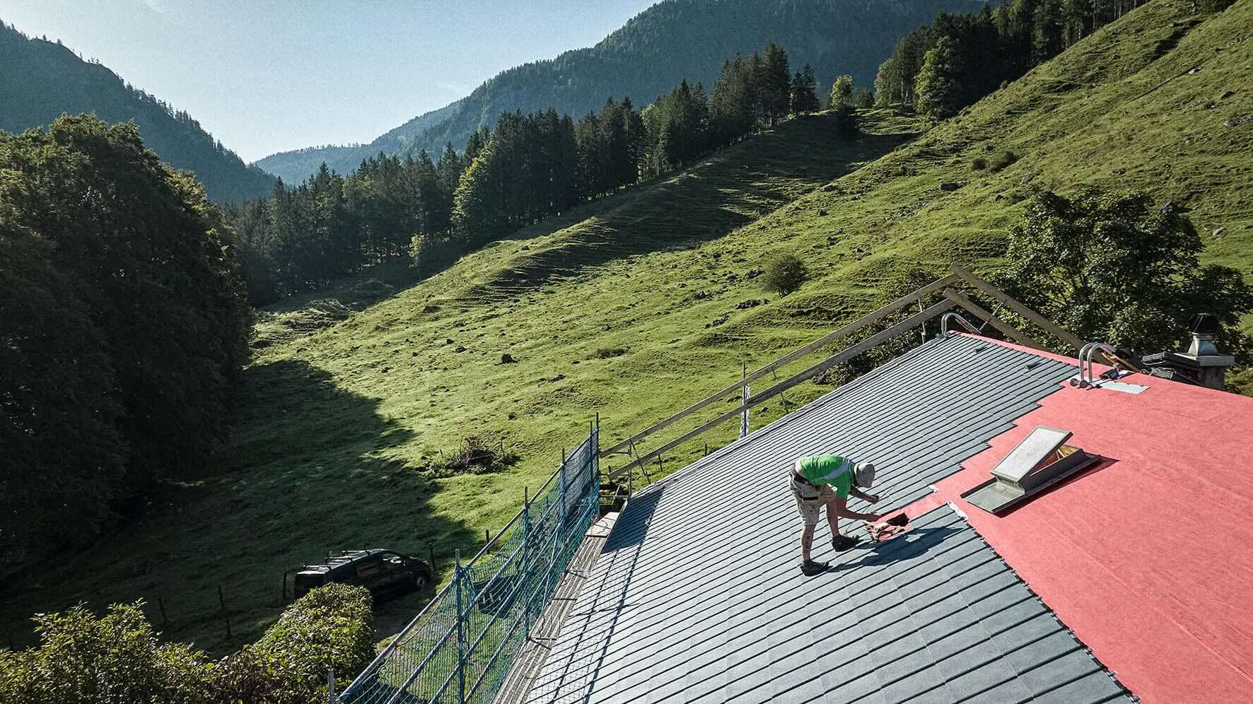 Craftsman roofing a pitched roof with grey tiles in a green mountain landscape.