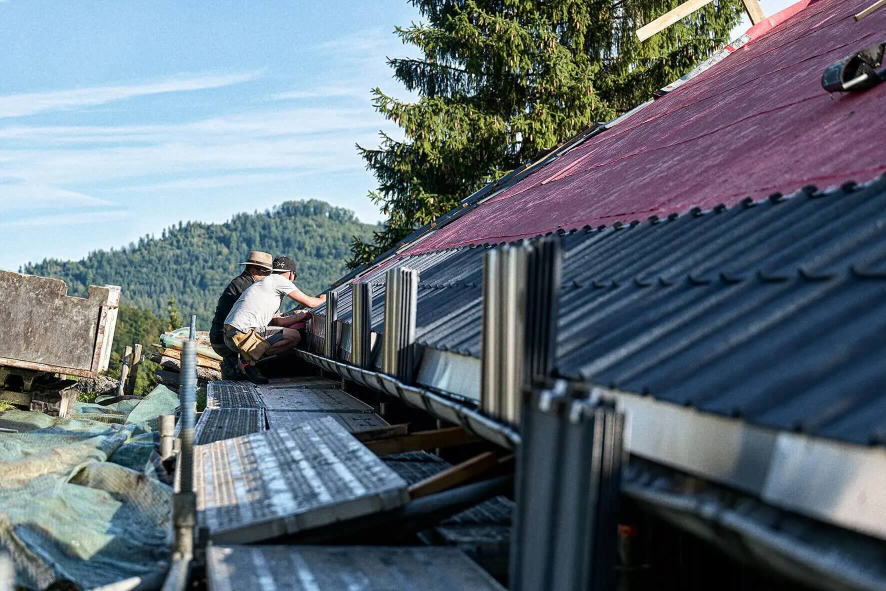Two men working on scaffolding near a roof in a rural, wooded area.