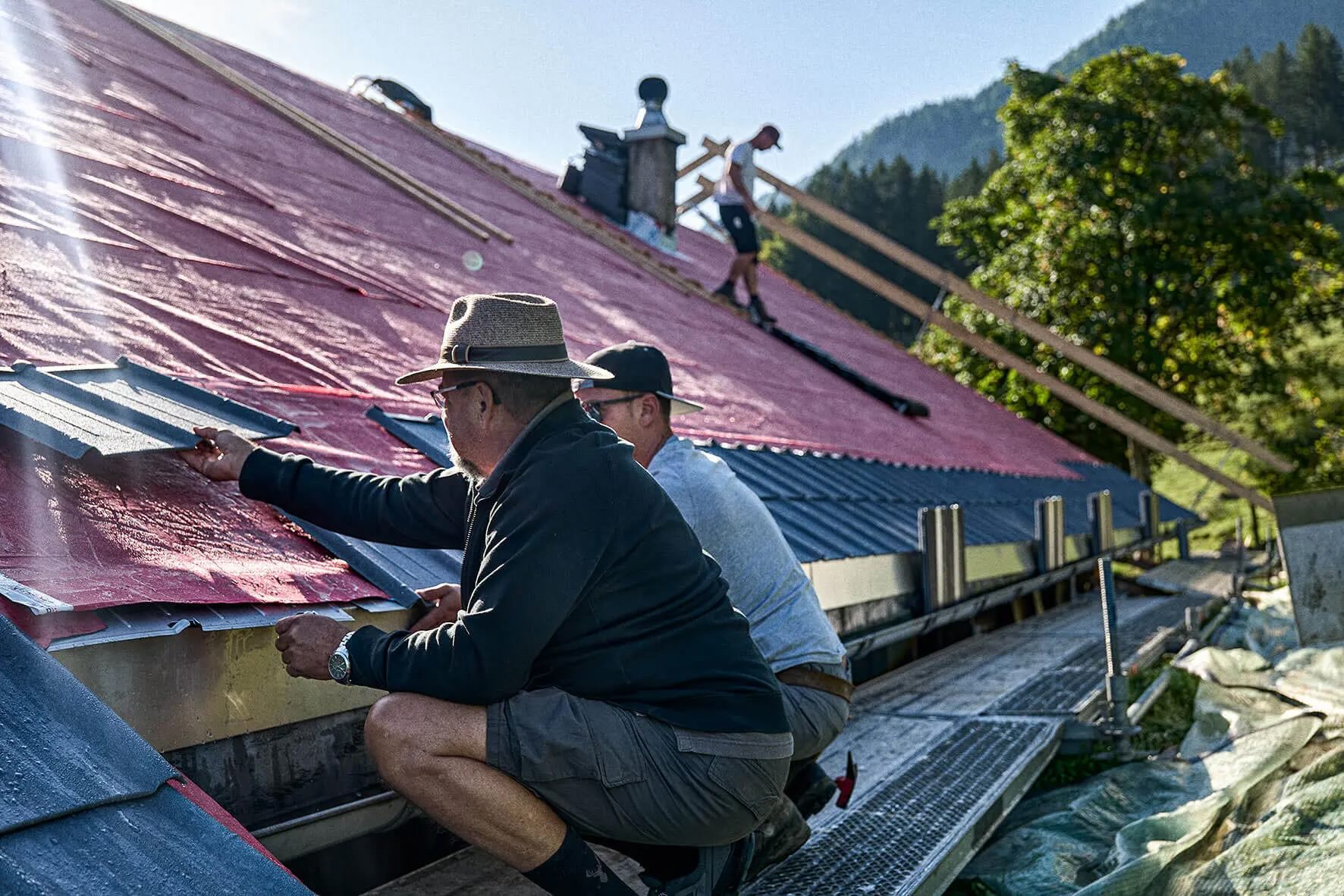 Three men installing roof tiles on a pitched roof in a sunny mountain landscape.