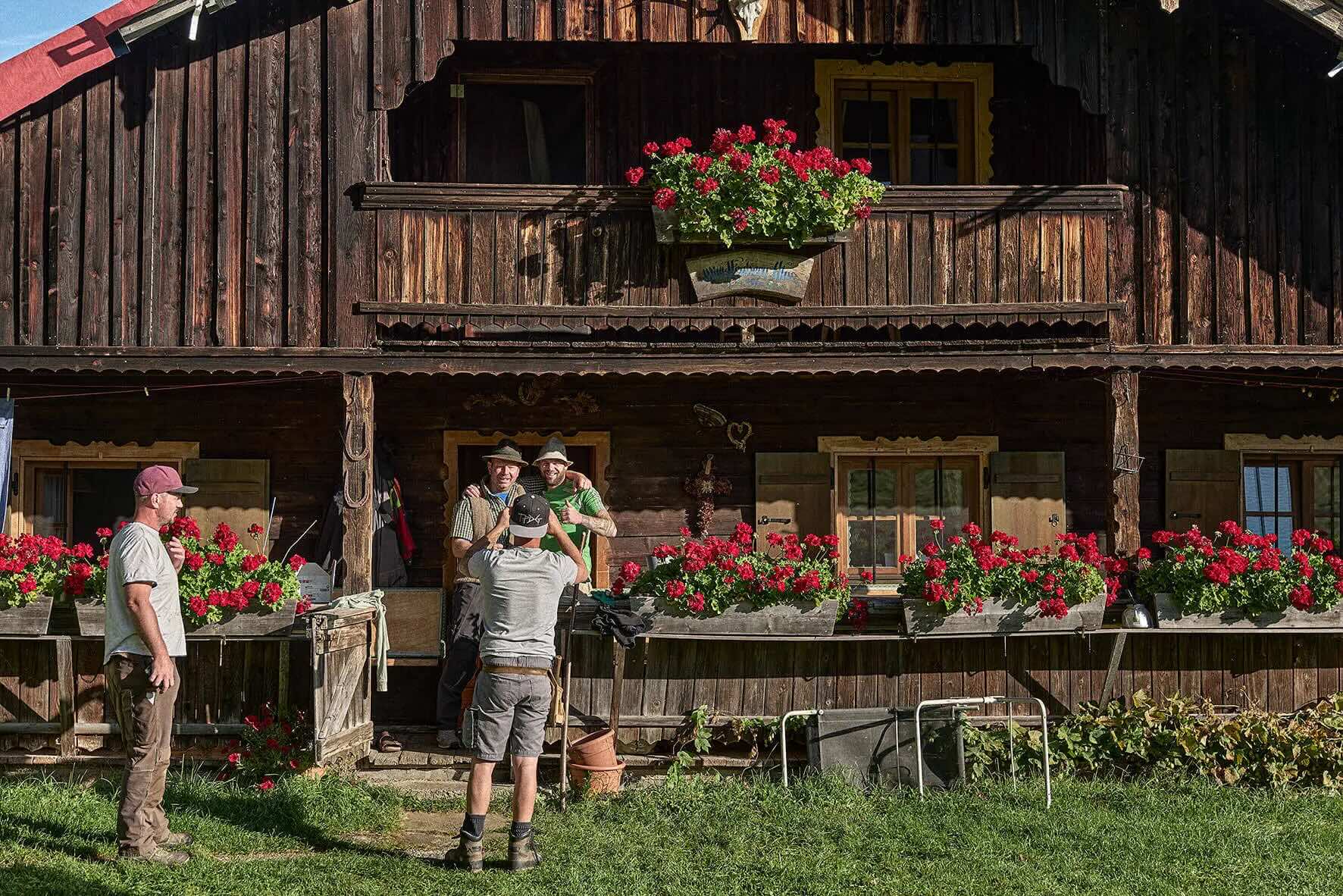 Three men in front of a rustic wooden hut with red flowers in window boxes, while one takes photos of the other two.