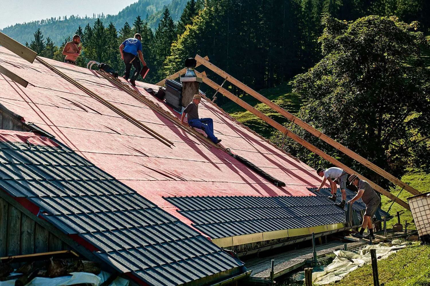 Several men are roofing a large roof with dark tiles in a mountainous, tree-filled landscape.