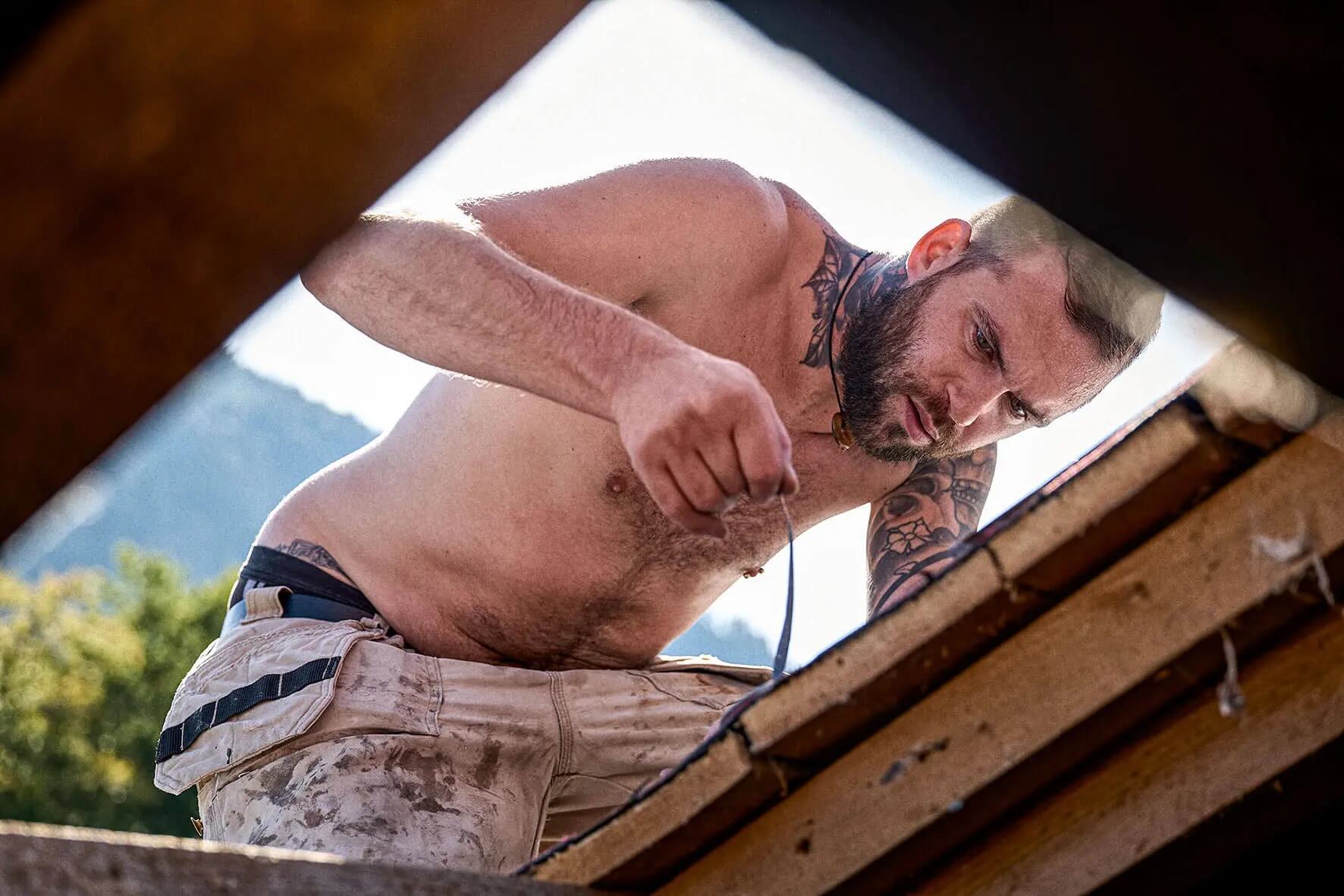 Shirtless man with tattoos working intently on a wooden roof outdoors.