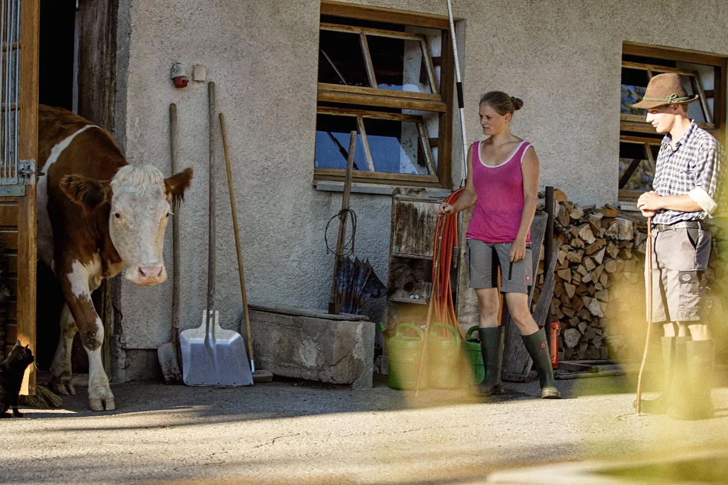 A woman and a man in rubber boots stand in front of a farm, while a cow emerges from the barn.