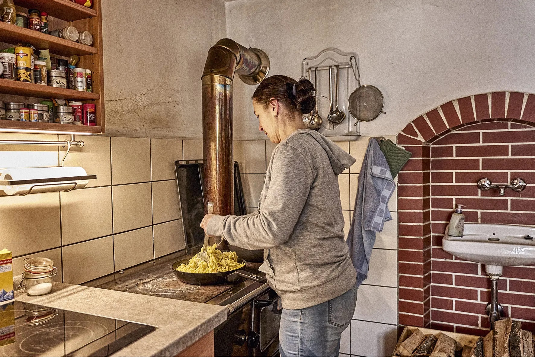 Frau in grauem Kapuzenpullover rührt Essen in einer Pfanne auf einem Herd in rustikaler Küche mit Holzofen und Gewürzregal.