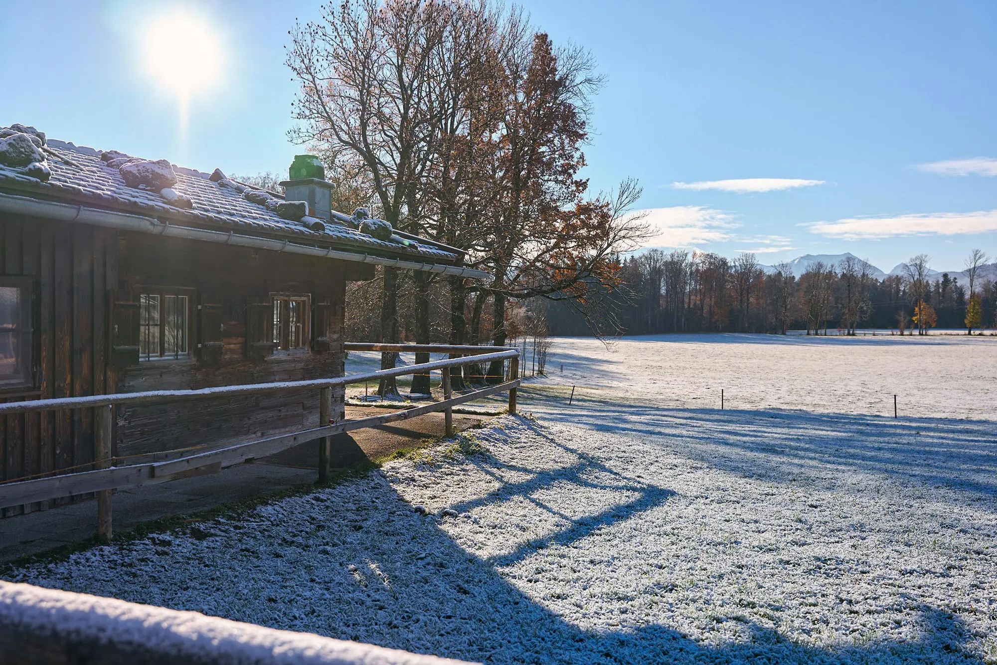 Holzhütte mit schneebedecktem Dach neben einem frostigen Feld unter klarem Himmel und strahlender Sonne.