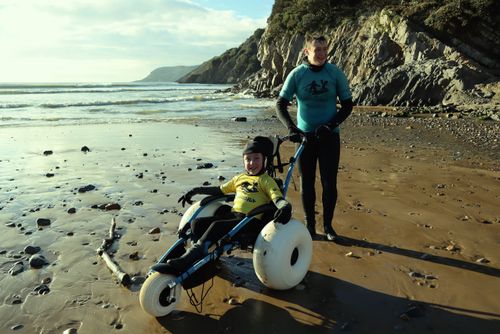 A boy in a beach wheelchair smiling post-surf with his surf instructor
