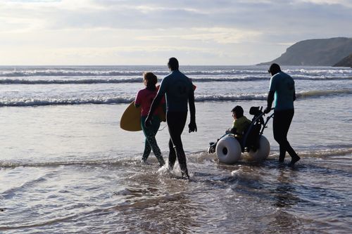 A young child in a beech wheelchair entering the sea with two instructors and a volunteer