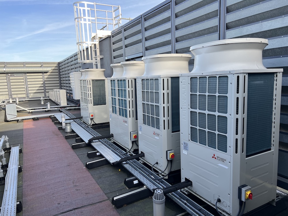 Row of Mitsubishi Electric rooftop HVAC units installed beside a metal safety railing and noise barrier on a building.