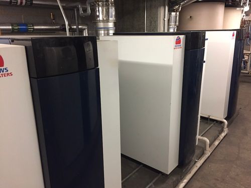 Row of white Andrews Water Heaters units installed indoors with visible piping and ventilation ducts above.