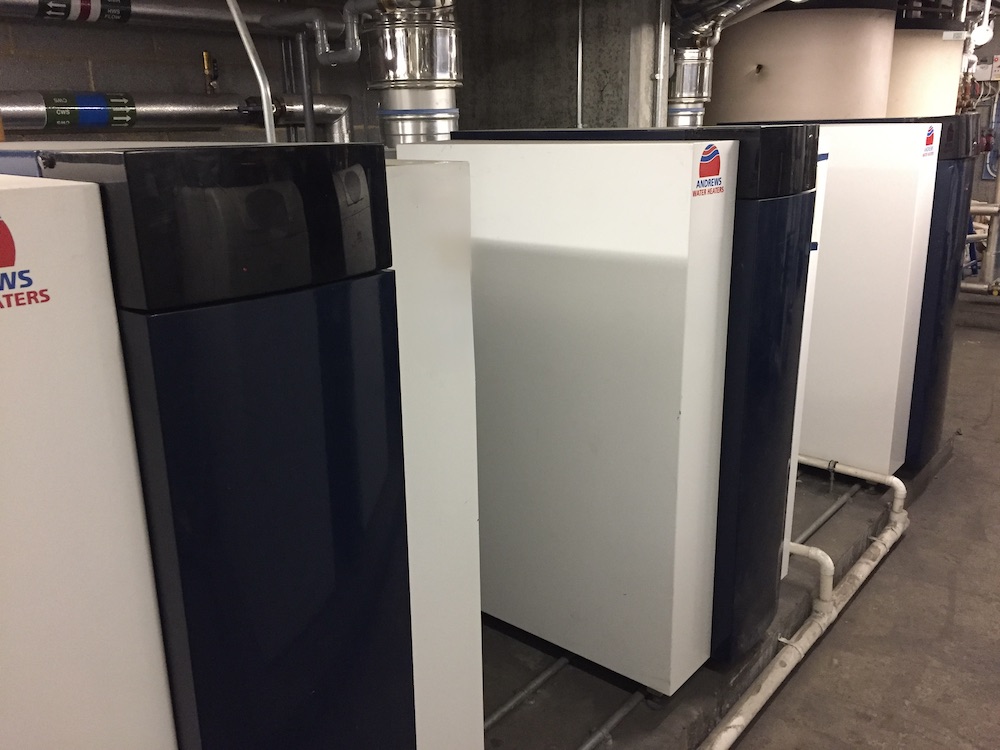 Row of white Andrews Water Heaters units installed indoors with visible piping and ventilation ducts above.