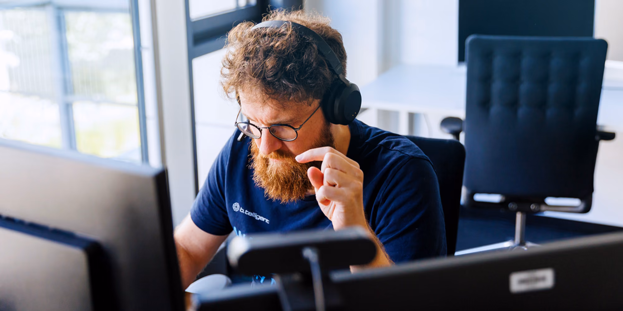 Mann mit Bart und Kopfhörern arbeitet konzentriert an Computern in einem Büro.