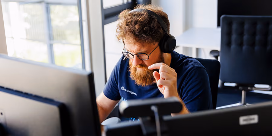 Mann mit Bart und Kopfhörern arbeitet konzentriert an Computern in einem Büro.