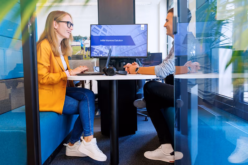 Two people in a modern office talking across a table with a computer screen showing MRM Marmind Solution.