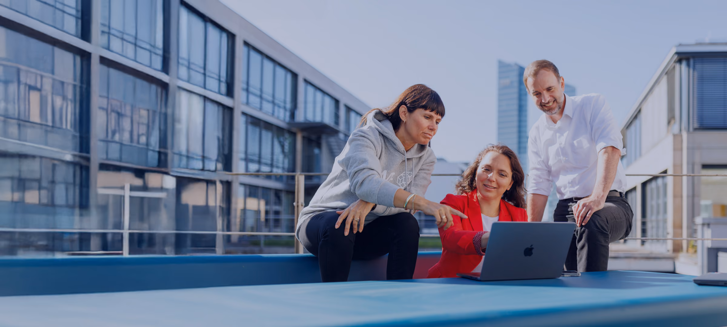 Drei Menschen im Freien sehen zusammen auf einen Laptop, eine Frau zeigt auf den Bildschirm.