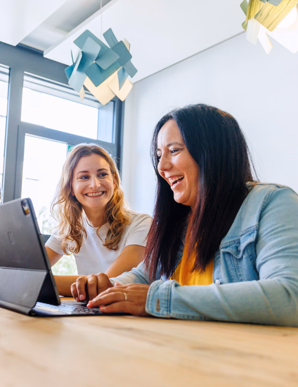 Zwei Frauen arbeiten lachend gemeinsam an einem Tablet in einem hellen Büro.