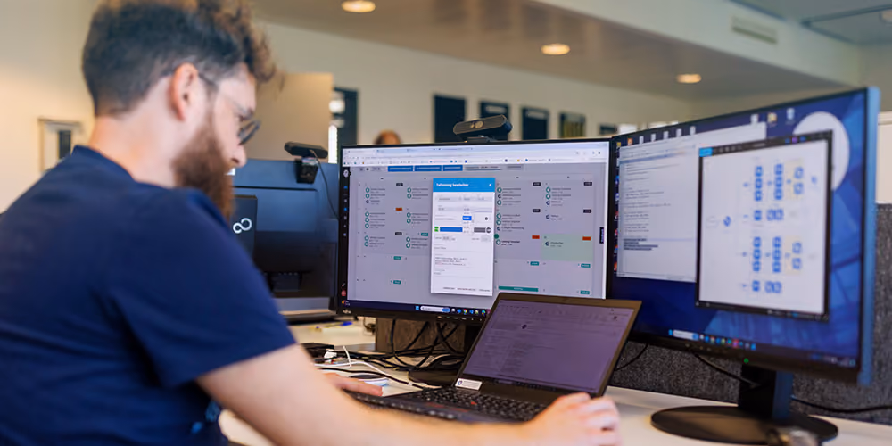 Mann sitzt vor drei Bildschirmen, die Softwareoberflächen mit Daten, Diagrammen und Einstellungsfenstern zeigen, in einem modernen Büro.
