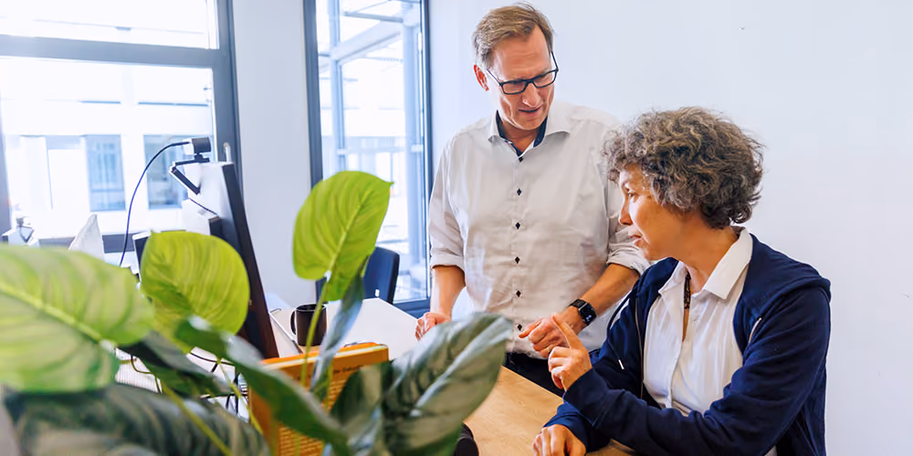 Zwei Menschen in einem modernen Büro mit großen Fenstern. Eine Frau sitzt an einem Schreibtisch und spricht mit einem Mann, der neben ihr steht und zuhört.