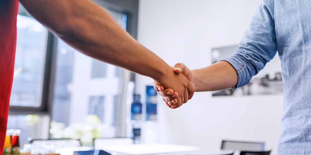 Zwei Personen geben sich die Hand in einem hellen Büro mit Fenstern im Hintergrund.