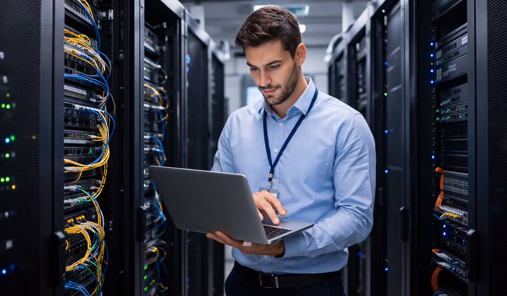 Man in a blue shirt working on a laptop inside a server room with network cables and racks.