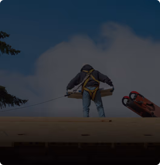 Construction worker wearing a safety harness carrying a plank on a rooftop with blue sky and clouds in the background.