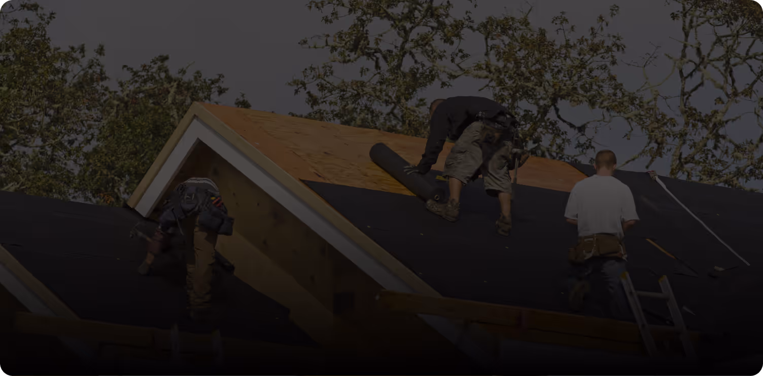 Three workers installing roofing felt on a house roof with trees in the background.