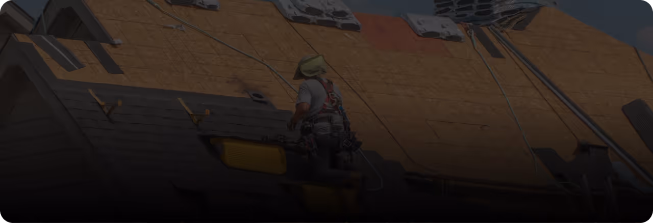 Construction worker wearing a safety harness and green hat working on the roof of a building under construction.