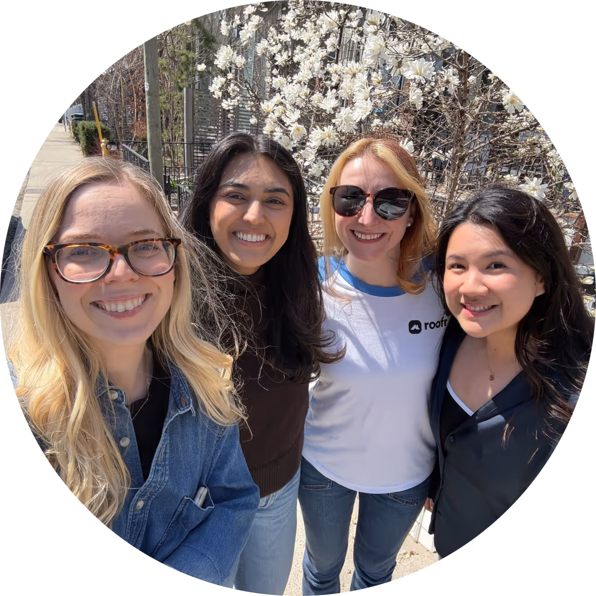 Four smiling women standing outdoors in front of a blooming white magnolia tree on a sunny day.