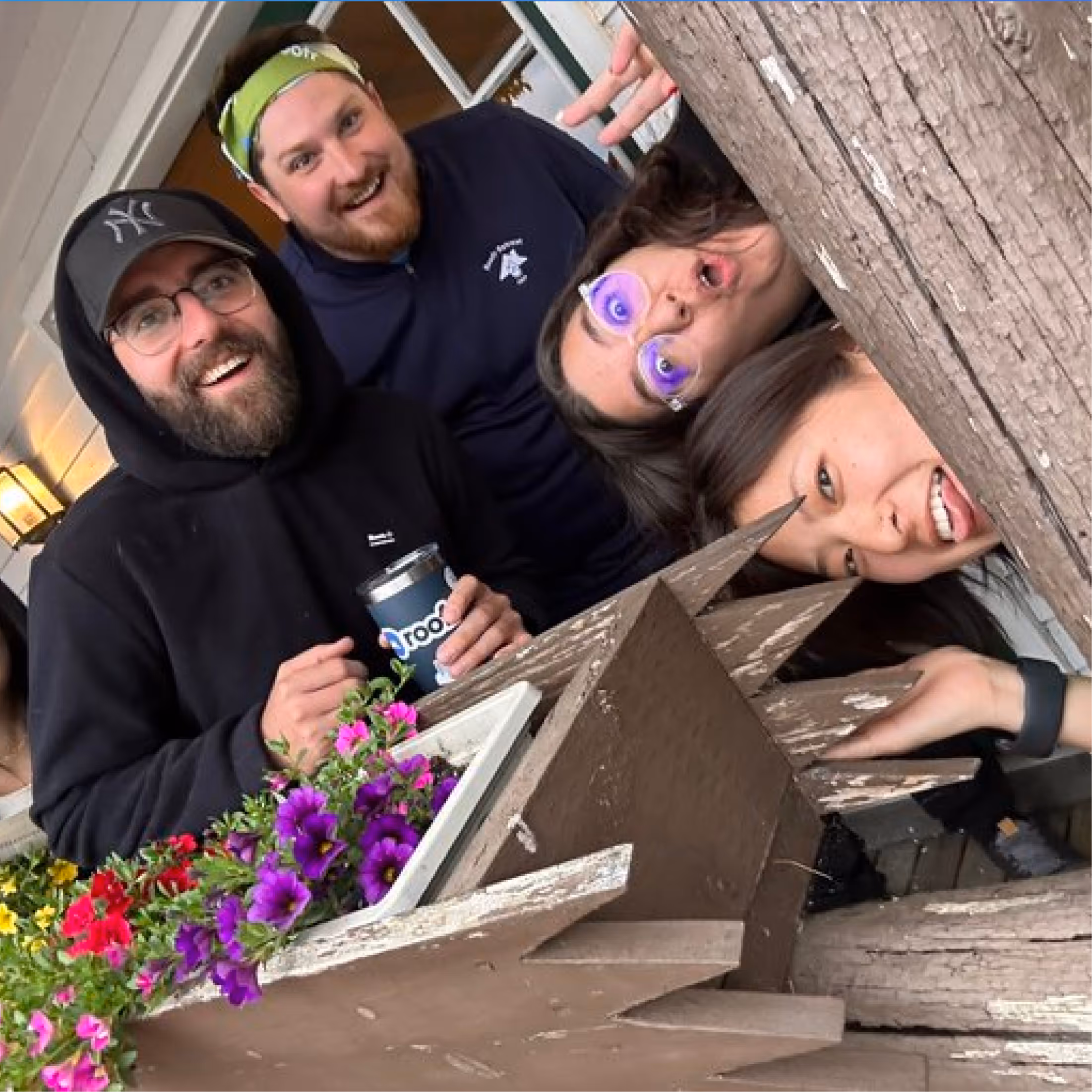 Four smiling friends leaning around a wooden planter box with colorful flowers on a porch.