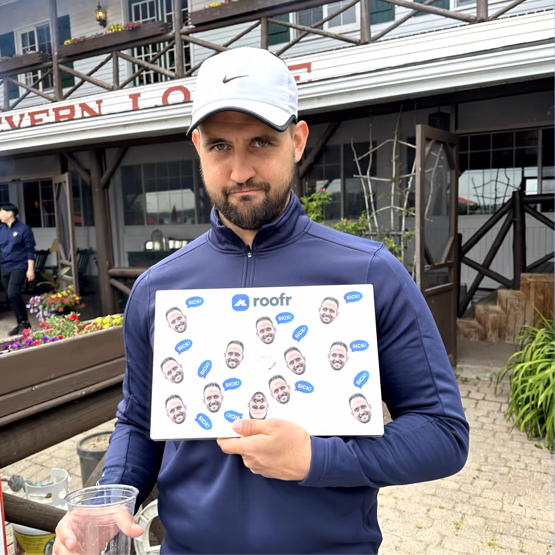 Man wearing a white Nike cap and navy jacket holding a laptop covered with stickers of a man's face and the word 'SICK!' in blue speech bubbles.