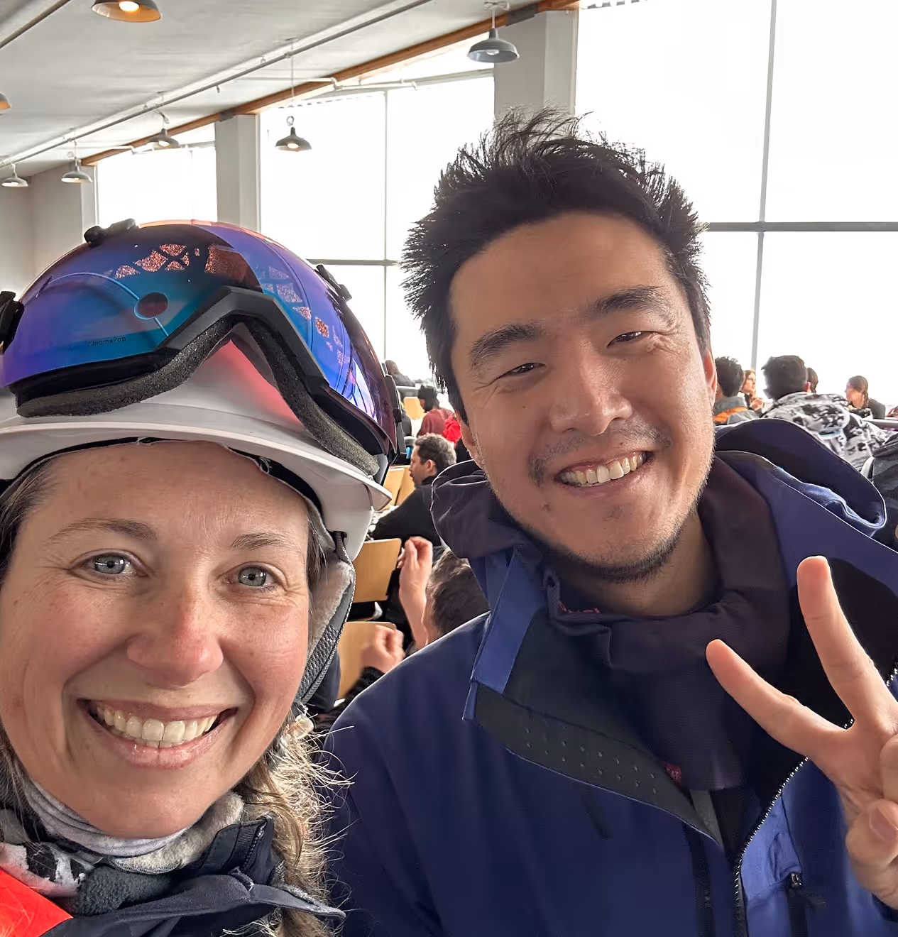 Smiling woman wearing a white helmet with blue ski goggles and a man in a blue jacket making a peace sign inside a busy room with large windows.