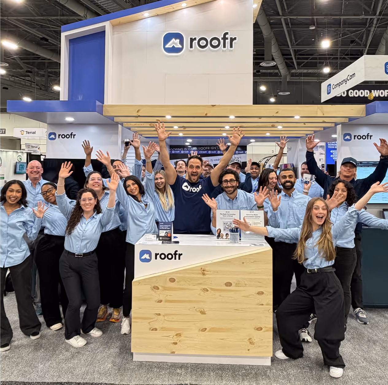 Group of roofr team members smiling and raising their hands in front of a roofr booth at an event.