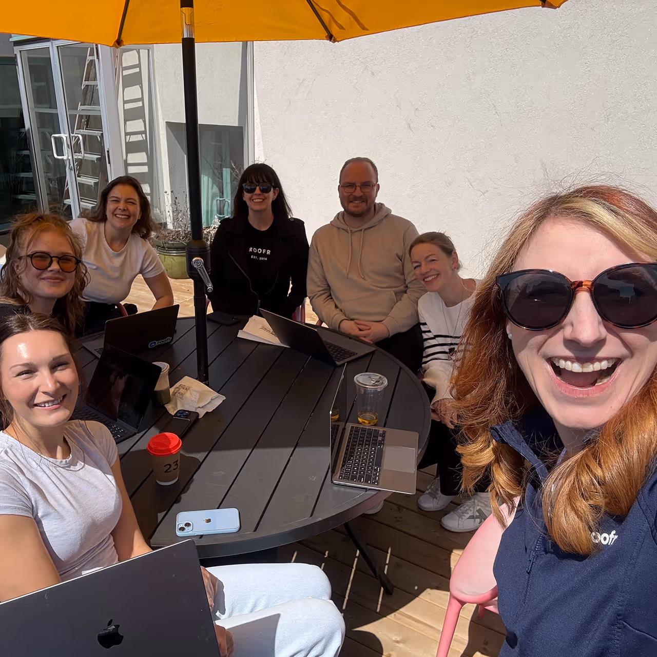 Seven smiling people sitting around an outdoor table with laptops under a yellow umbrella on a sunny day.