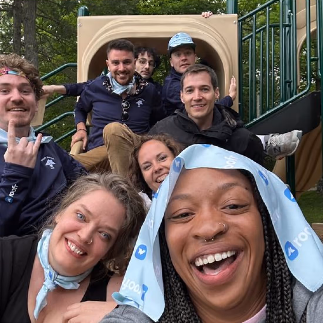 Group of eight smiling people posing for a selfie on playground equipment outdoors.