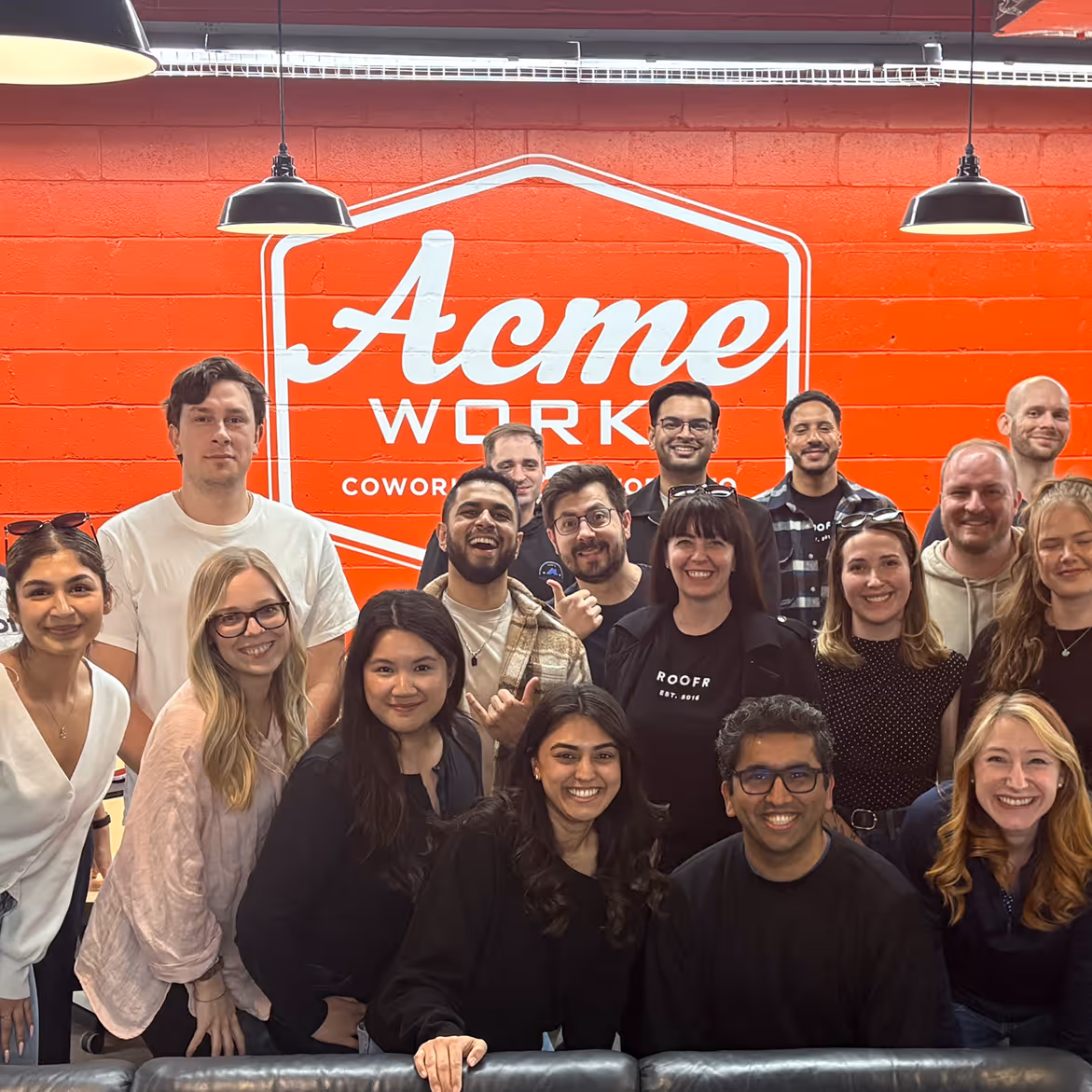 Group of smiling young adults posing inside a coworking space with a bright orange wall featuring a large Acme Work sign.