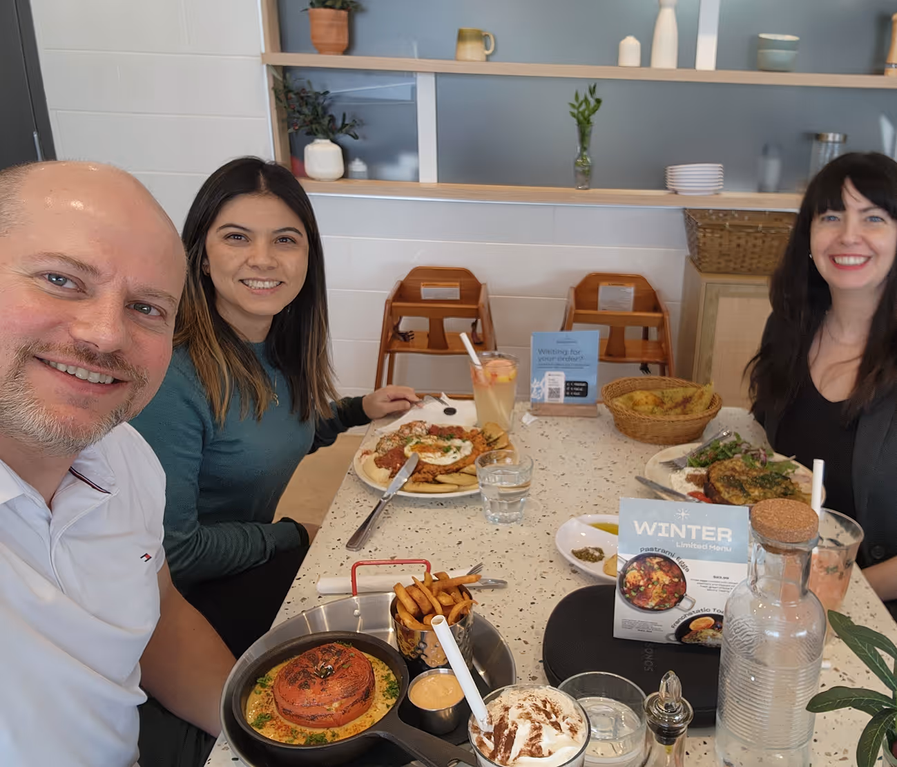Three people smiling and sitting at a table with various dishes including fries, salad, and drinks in a restaurant setting.