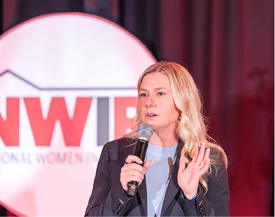 Woman with blonde hair speaking into microphone on stage with NWIR backdrop behind her.