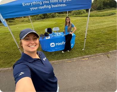 Two women smiling at a roofing business booth with a blue canopy and tablecloth outdoors.