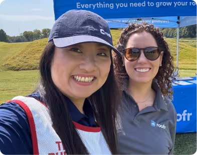 Two smiling women outdoors, one wearing a blue cap and white vest, the other in sunglasses and a gray shirt.
