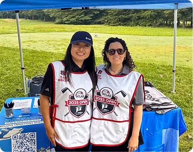 Two women smiling under a canopy wearing 2025 Open event vests at a golf course registration table.