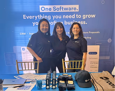 Three women in navy shirts behind a table with promotional items at a business booth.